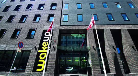Gadebillede af en moderne murstensbygning med store vinduer. To flag er foran, og på et skilt står "vågning" ved indgangen. Et "parkeringsforbud"-skilt er også synligt på fortovet.