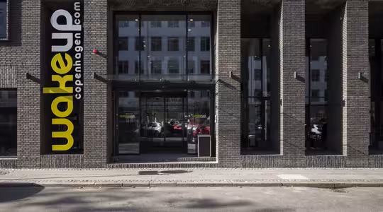 Billedet viser en moderne bygning med et ydre i mørk mursten. På et stort lodret skilt står "wake up copenhagen.com" med gule og hvide bogstaver. Glasdøre fører indenfor og afspejler nærliggende bygninger og parkerede biler.