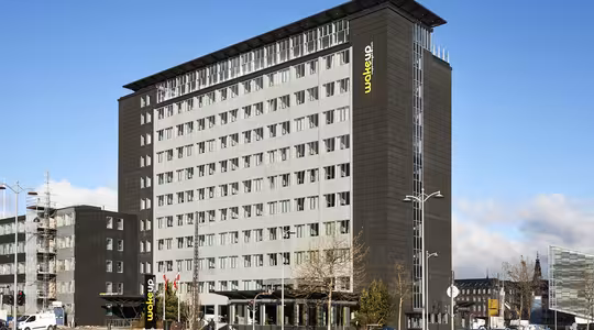 En moderne hotelbygning i flere etager med en grå facade og store vinduer. "Wakeup" står der med gult på siden. Forgrunden viser en gade med biler, cykler og spredte fodgængere under en delvist overskyet himmel.