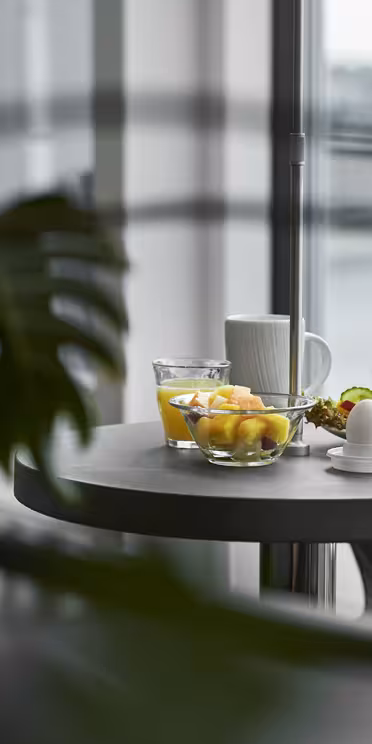 Et bord ved et stort vindue er dækket med en skål frugt, et glas vand, en kop kaffe og en tallerken med en sandwich og kogt æg. Grønne blade indrammer delvist forgrunden med en udendørs udsigt ud over vinduet.