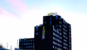 En moderne etagebyggeri med mørkt ydre og stor logotekst "wakeup" i grønt og hvidt på den øverste facade. Himlen er klar med en lys rosa nuance. Bygningen har flere vinduer og er omgivet af et lavt hegn.