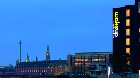 Bybillede i skumringen med en forlystelsespark i det fjerne og et hotel med "vågn op"-skilte. Tage og historiske bygninger er synlige under en mørkere blå himmel.