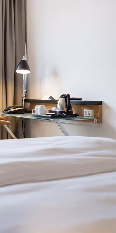 Hotelværelse med en pæn opredt seng, et væghængt tv og et skrivebord ved vinduet. Skrivebordet har en moderne lampe, en stol og en kedel med kopper. Gardiner dækker en del af vinduet, og træer kan ses udenfor.