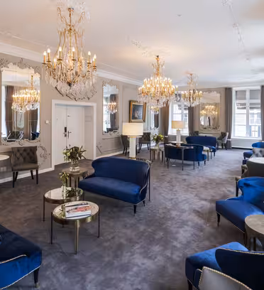 Elegant møbleret lounge med siddepladser i blåt fløjl, udsmykkede lysekroner og store spejle på væggene. Små runde borde med magasiner og dekorative vaser er placeret i hele rummet, som har gråt tæppe og høje vinduer.