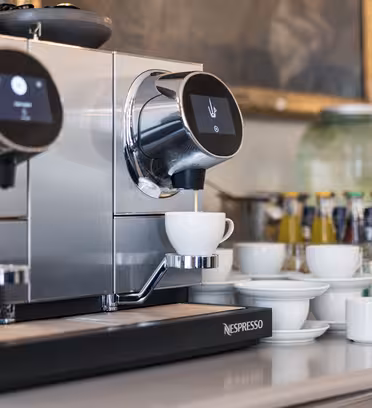En elegant espressomaskine med to dispensere laver kaffe til en hvid kop. I nærheden er stablede kopper, underkopper og glas. I baggrunden står flasker og krukker på en disk.