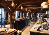 Et rummeligt, varmt oplyst restaurantinteriør med træbjælker og en buffetopstilling. Rustik indretning suppleres med hængende pærer. Der er arrangeret borde med mad, hvilket skaber en indbydende spisestemning.