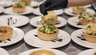 En person, der bærer en sort handske, pynter små tarteletter fyldt med blandet grønt og hakkede ingredienser på hvide tallerkener, arrangeret i rækker. Tallerkenerne har dekorative drys af sauce ved siden af tarteletterne.