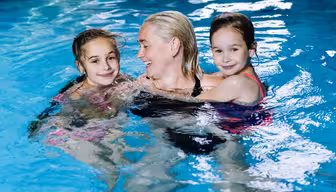 En kvinde smiler i en swimmingpool med to unge piger. De nyder alle deres tid i det klare blå vand, hvor pigerne krammer kvinden. Scenen er munter og afslappet.