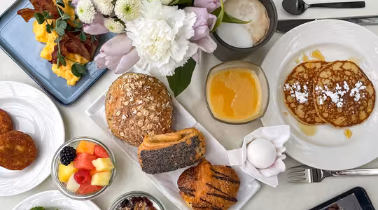 Et morgenmadspålæg med røræg, sprød bacon, diverse kager, pandekager med pulveriseret sukker, frugt, yoghurt med nødder, et kogt æg og en frugtdrik. En buket blomster, en cappuccino og en smartphone er også synlige på bordet.