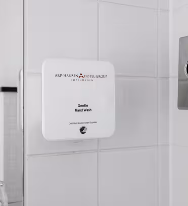 Væghængt sæbedispenser i badeværelse med Arp-Hansen Hotel Groups logo. Ved siden af er der en vævsholder. Væggene er flisebelagt i hvidt, og to glaskopper er delvist synlige på en hylde under.