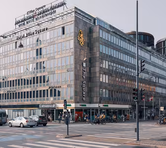 Travlt vejkryds i en by med en stor hjørnebygning med skiltning til "Hotel The Square". Cykler er parkeret udenfor, og fodgængere og biler er synlige. Arkitekturen omfatter rektangulære vinduer og urbane modernistiske designelementer.