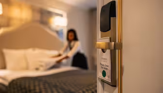 Hotelværelse med åben dør med fokus på et "Please clean the room"-skilt. Sløret baggrund viser en person, der laver sengen. Værelset har en hyggelig stemning med blød belysning og elegant indretning.