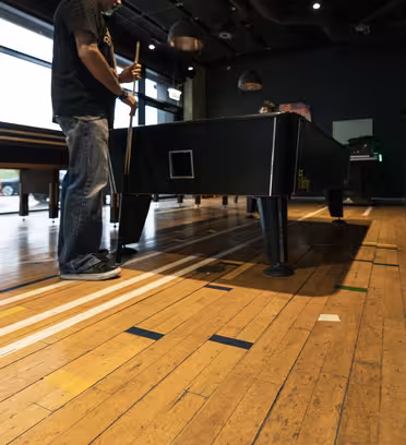 En person står ved et shuffleboard-bord i et moderne indendørs spillerum med trægulve og store vinduer. De holder en stikordspind iført en mørk skjorte og jeans. Rummet er godt oplyst med loftslamper og viser reflekser på det skinnende gulv.