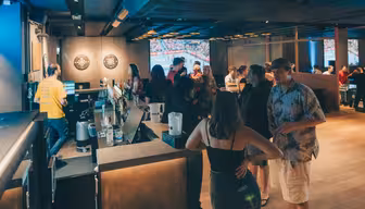 En svagt oplyst bar med folk, der hygger sig nær disken. En kvinde i en sort top står i forgrunden, mens en mand i en blomstret skjorte og shorts er i nærheden. Bartendere serverer drinks, og dartskiver er synlige på væggen.