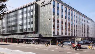 En moderne hjørnebygning med store vinduer huser Imperial-hotellet og restauranten LApetito. Folk går og cykler på gaden foran, og biler er parkeret langs vejkanten. Det er en solskinsdag.