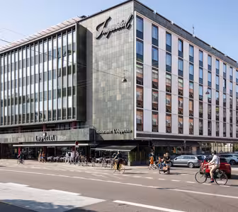 En moderne hjørnebygning med store vinduer huser Imperial-hotellet og restauranten LApetito. Folk går og cykler på gaden foran, og biler er parkeret langs vejkanten. Det er en solskinsdag.