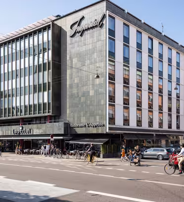 En moderne hjørnebygning med store vinduer huser Imperial-hotellet og restauranten LApetito. Folk går og cykler på gaden foran, og biler er parkeret langs vejkanten. Det er en solskinsdag.