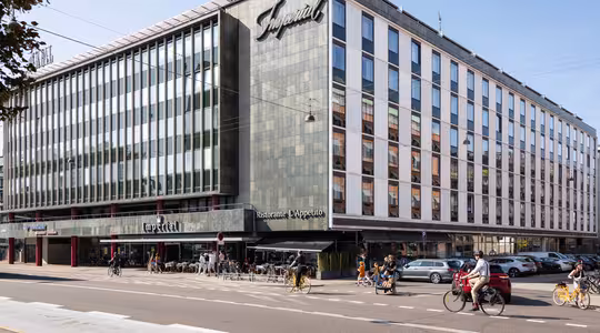 En moderne hjørnebygning med store vinduer huser Imperial-hotellet og restauranten LApetito. Folk går og cykler på gaden foran, og biler er parkeret langs vejkanten. Det er en solskinsdag.