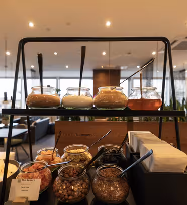 En buffetudstilling med et udvalg af saucer og toppings i glaskrukker og skåle med serveringsskeer. Opsætningen er inde i en moderne restaurant med varm belysning og planter i baggrunden.