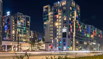 Natscene af et moderne hotelkompleks med flere bygninger. Strukturerne er stærkt oplyste og byder på farverigt oplyste vinduer. En vej med kørende billygter er i forgrunden, og gadelygter langs vejen.