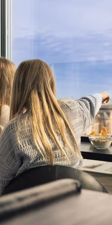To personer sad indendørs ved et vindue, den ene peger ud mod et bybillede med hustage og spir. En skål med snacks står på bordet. Scenen antyder en afslappet atmosfære med panoramaudsigt.