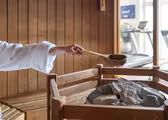 En person i en hvid morgenkåbe hælder vand fra en slev på varme saunasten inde i et saunarum af træ. Fitnessudstyr er synligt i baggrunden.