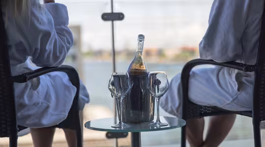 To personer i badekåber sidder på stole ud mod et glasrækværk med udsigt over bybilledet. Et lille bord rummer en spand med en flaske vin og to glas. Scenen antyder afslapning og fritid.