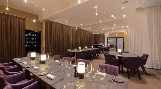 Interiør i en moderne, elegant restaurant med lilla stole og brune borde med glas og tallerkener. Varm, dæmpet belysning fra hængende pærer skaber en hyggelig atmosfære. Brune og hvide gardiner opdeler rummet.