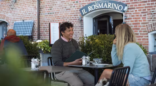 To personer sidder ved et udendørs bord på en restaurant ved navn Il Rosmarino, der ligger foran en murstensvæg. Manden holder en menu og smiler til kvinden, der har langt hår og hviler hagen på hånden.