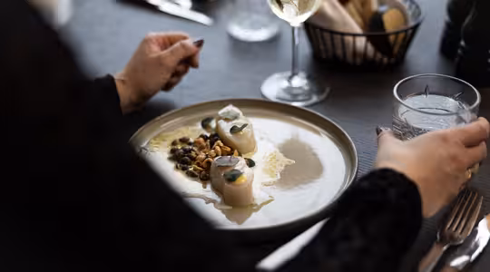 En person holder et glas vand ved siden af en tallerken gourmetmad, inklusive kammuslinger og pynt, på et bord. Et glas hvidvin og en kurv med brød er i baggrunden. Indstillingen ser ud til at være en fin restaurant.