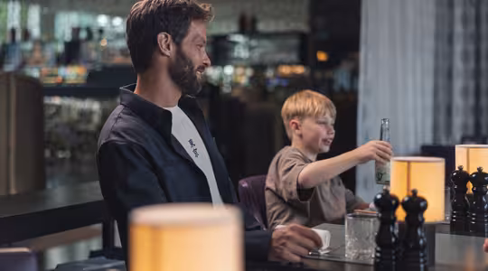 En mand og et barn sidder ved et svagt oplyst bord i en restaurant. Manden kigger mod barnet, som smiler og holder en flaske. Bordet har tændte lamper og sorte peberkværne. Baggrunden viser et sløret restaurantinteriør.