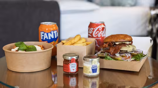 Et glasbord med fastfood: en kasse pommes frites, en burger med salat, ost og bacon, en skål pasta pyntet med basilikum, dåser Fanta og Coca-Cola og to små krukker med krydderier. Baggrunden viser en sløret seng og puder.