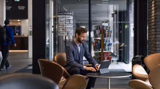 En mand i jakkesæt sidder i en moderne kontorlobby og arbejder på en bærbar computer ved et rundt bord. Området er stilfuldt indrettet med brune stole og har brochurestander i baggrunden. En anden person går væk i baggrunden.