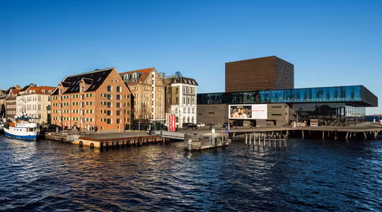 Udsigt ved vandet med en række historiske murstensbygninger og en moderne glasstruktur med et fladt tag, der strækker sig over vandet. Bådene ligger til kaj langs træmolerne under en klar blå himmel.