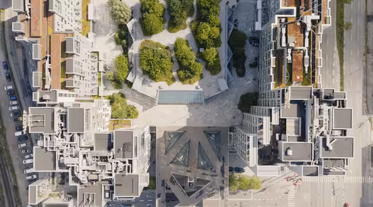 Luftfoto af et byområde med moderne bygninger omkring en central plads fyldt med træer. Gader og stier er synlige, med varieret arkitektonisk design i bygningerne. Billedet fanger en blanding af grønne områder og byudvikling.