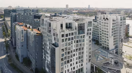 Luftfoto af moderne højhuse med glasfacader og tagterrasser i et byområde. Tilstødende strukturer omfatter en mørk bygning med orange tage og en glaspyramideindgang. Skyline og flod er synlige i baggrunden.