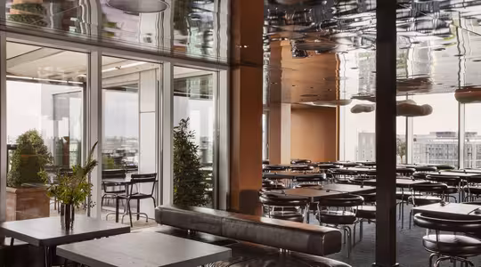 Moderne indendørs cafe med mørke lædersæder, træborde og gulv til loft-vinduer. Reflekser er synlige på det skinnende loft. Udendørs terrasse med planter og udsigt til bybilledet i baggrunden.