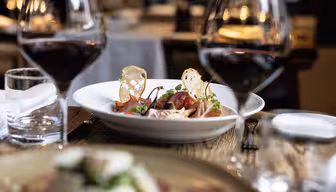 Et spisebord med en tallerken gourmetmad, med skåret kød og pynt, mellem to glas rødvin. Baggrunden viser slørede restaurantsæder, hvilket skaber en hyggelig atmosfære.