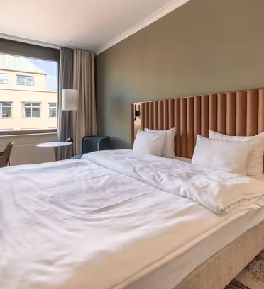 Et moderne hotelværelse har en stor, pænt redt seng med hvidt sengetøj og en polstret sengegavl. Et skrivebord med en stol er nær vinduet, og et lille bord med en stol er på den modsatte side. Væggene er malet grønne.