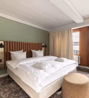 Et moderne hotelværelse har en stor seng med hvidt sengetøj og en træsengegavl mod en salviegrøn væg. Der er en beige skammel ved fodenden af sengen, og store vinduer med tunge gardiner har udsigt til et industrielt bybillede.