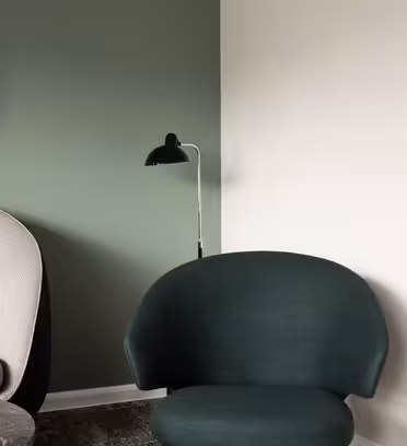 Et moderne interiør har en beige lænestol og en mørkegrøn stol. Et sort, rundt spejl hænger på en grøn væg, og en sort gulvlampe står ved siden af. En hvid væg står i kontrast til hjørnet af rummet.