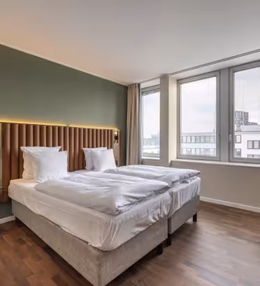 Hotelværelse med to pænt opredte senge mod en grøn accentvæg. Værelset har store vinduer med udsigt over byen, et lille rundt bord og en mørkegrøn lænestol. Naturligt lys fylder det minimalistiske rum og fremhæver trægulvet.