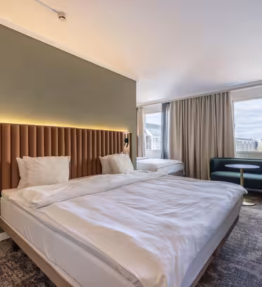 Hotelværelse med to senge, moderne indretning og jordfarver. Inkluderer en polstret sengegavl, grøn accentvæg, store vinduer med udsigt over byen, grøn sofa, rundt bord og stole. Tæppebelagte gulve og gardiner forbedrer rummenes hyggelige stemning.