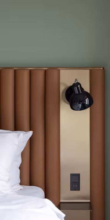 Moderne soveværelse med en grøn væg, en brun polstret sengegavl og en pæn opredt seng med hvide puder og lagner. En sort væglampe og en stikkontakt er monteret på et metalpanel ved siden af sengen. Beige gardin til højre.