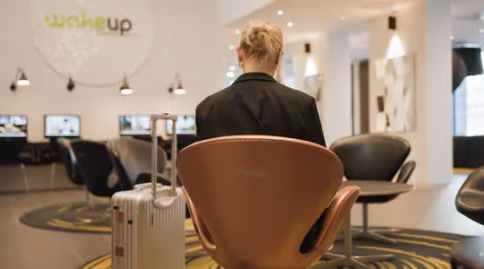 En person med tilbagebundet hår sidder i et moderne venteområde på en rund stol med Wakeup trykt på. De vender væk, med en sølvkuffert ved siden af sig. Rummet har slanke, sorte stole og computere i baggrunden.
