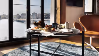 En morgenmad med brød, kager, frugt og drikkevarer er dækket op på et rundt bord ved store vinduer med udsigt over et bybillede ved vandet, hvor naturligt lys strømmer ind i rummet.
