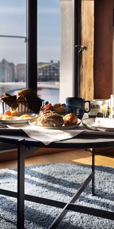 En morgenmad med brød, kager, frugt og drikkevarer er dækket op på et rundt bord ved store vinduer med udsigt over et bybillede ved vandet, hvor naturligt lys strømmer ind i rummet.