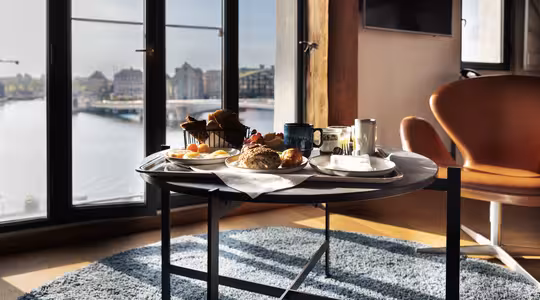 En morgenmad med brød, kager, frugt og drikkevarer er dækket op på et rundt bord ved store vinduer med udsigt over et bybillede ved vandet, hvor naturligt lys strømmer ind i rummet.