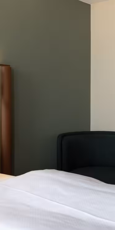 Et moderne hotelværelse har en seng med hvidt sengetøj og puder, en polstret brun hovedgærde, et lille rundt sidebord, en grøn polstret stol og en hængende cylindrisk lampe op mod tofarvede vægge.