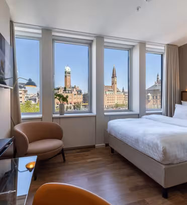 Et moderne hotelværelse med en stor seng, et tv, en brun lænestol, et skrivebord og tre store vinduer med udsigt over byen med historiske bygninger og tårne under en klar blå himmel.
